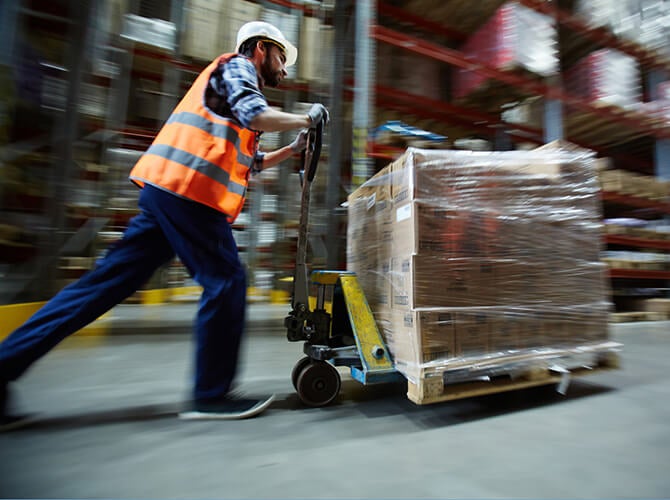 A warehouse worker in a high-visibility vest and helmet expertly maneuvers a pallet jack loaded with wrapped boxes of specialized freight. The background reveals tall shelves brimming with diverse items, highlighting the bustling energy of the storage area.