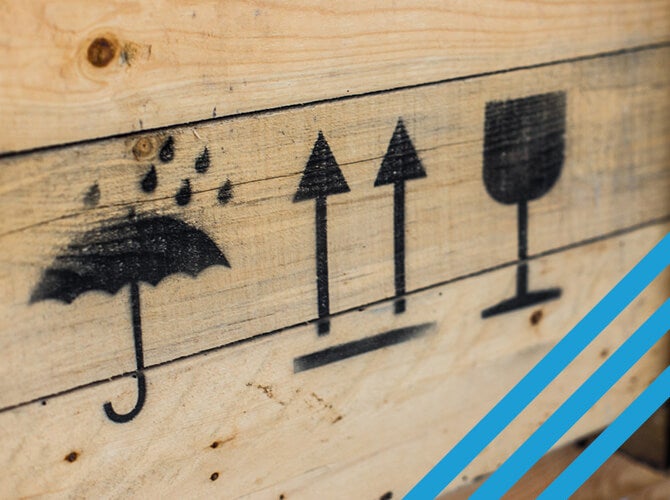 Symbols painted on a wooden crate for specialized freight: an umbrella with raindrops indicating keep dry, two upward arrows signifying this side up, and a wine glass symbol meaning fragile. Diagonal blue stripes adorn the bottom right corner.