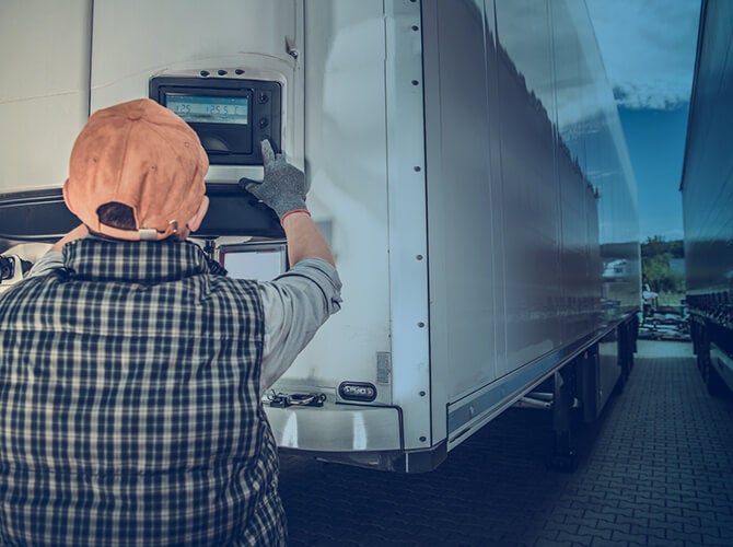 A person in a plaid shirt, cap, and glove expertly operates the control panel on a large refrigerated truck, specializing in freight. The vehicle is parked on a paved surface under a cloudy sky.