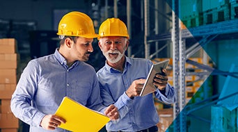 Two business partners in formal wear and with protective yellow helmets on heads standing in warehouse and comparing data. Younger holding folder while older one pointing at calculations on tablet.