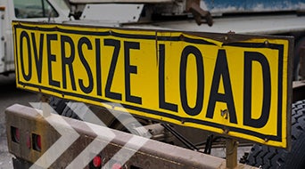 A bright yellow sign with black text reading OVERSIZE LOAD is mounted on the back of a truck. The background shows part of the truck and faint white arrow markings on the pavement.