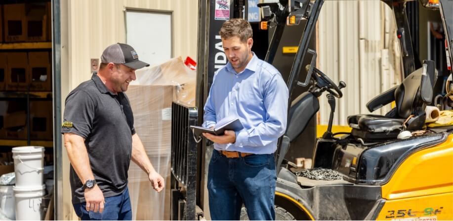 Two men stand next to a forklift outside a warehouse. One man, wearing a black shirt and cap, smiles while the other, in a blue shirt, holds a notebook and appears to discuss something with him.